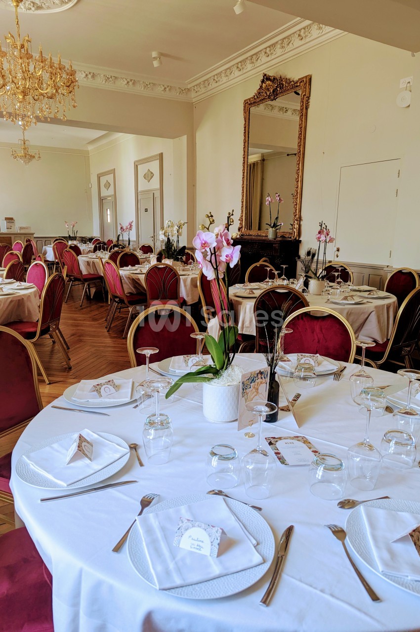 Salle de réception élégante avec tables rondes dressées, nappes blanches, chaises rouges et décorations florales.