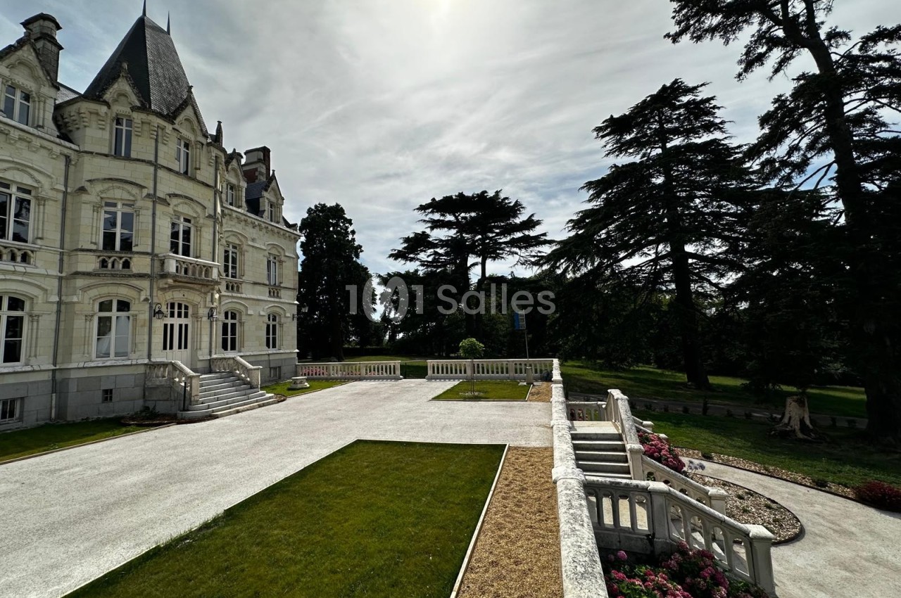 Façade d'un château avec terrasse en pierre, pelouses aménagées et arbres imposants sous un ciel partiellement nuageux.