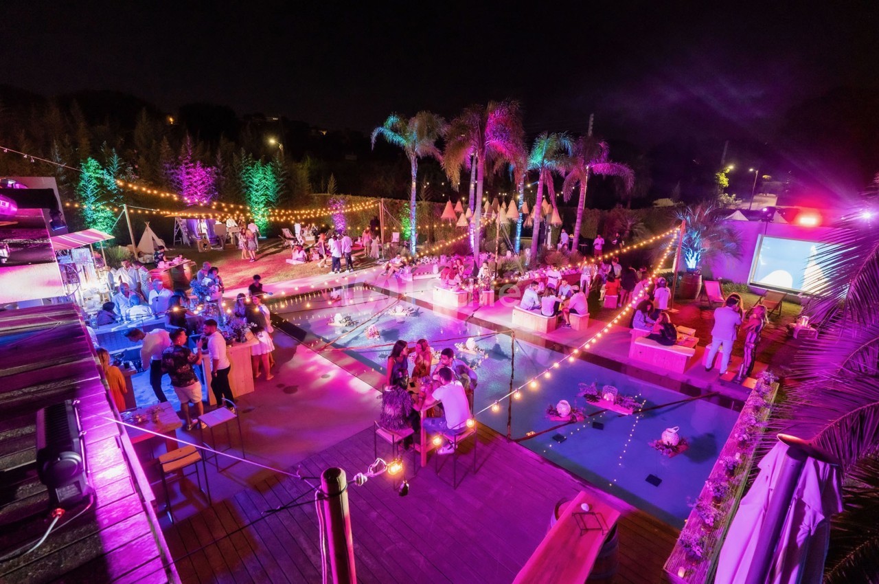 Vue nocturne d'une fête en plein air autour d'une piscine, éclairée par des guirlandes lumineuses et des projecteurs colorés.