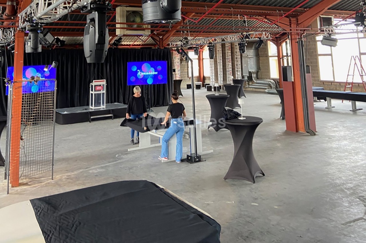 Deux personnes préparent une salle industrielle avec des tables hautes et des écrans pour un événement.