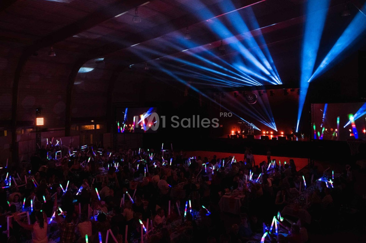 Salle sombre avec un public tenant des bâtons lumineux colorés, éclairée par des projecteurs et des faisceaux lumineux.