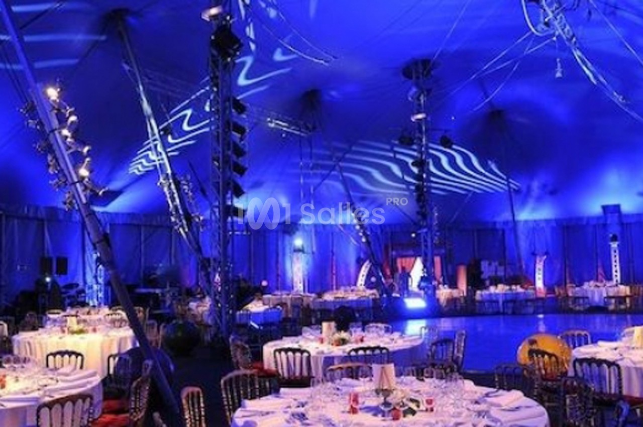 Salle de réception sous un chapiteau éclairé en bleu, avec des tables rondes dressées pour un événement.