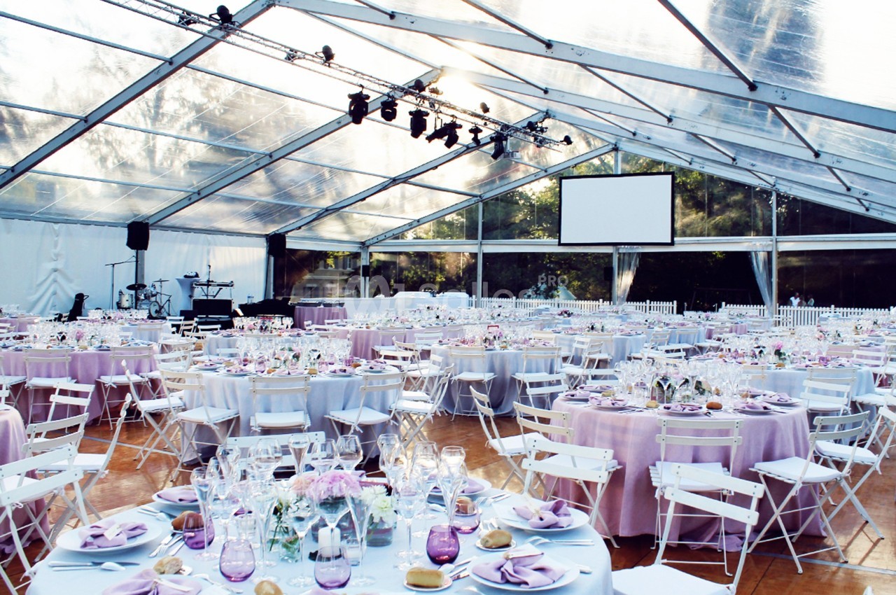 Salle de réception sous une tente transparente avec des tables rondes décorées de nappes violettes et des chaises blanches.