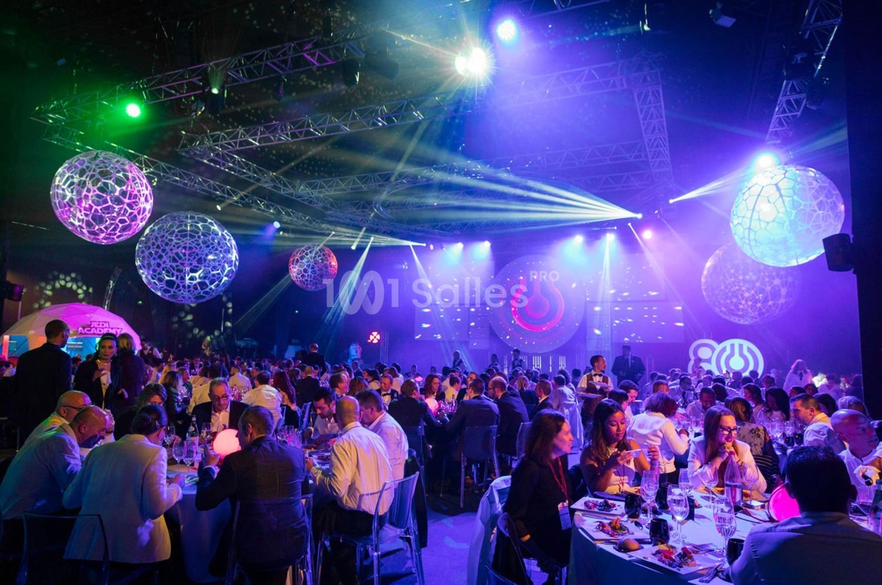 Salle de réception illuminée avec des invités assis à des tables, des décorations suspendues et des jeux de lumière colorés.
