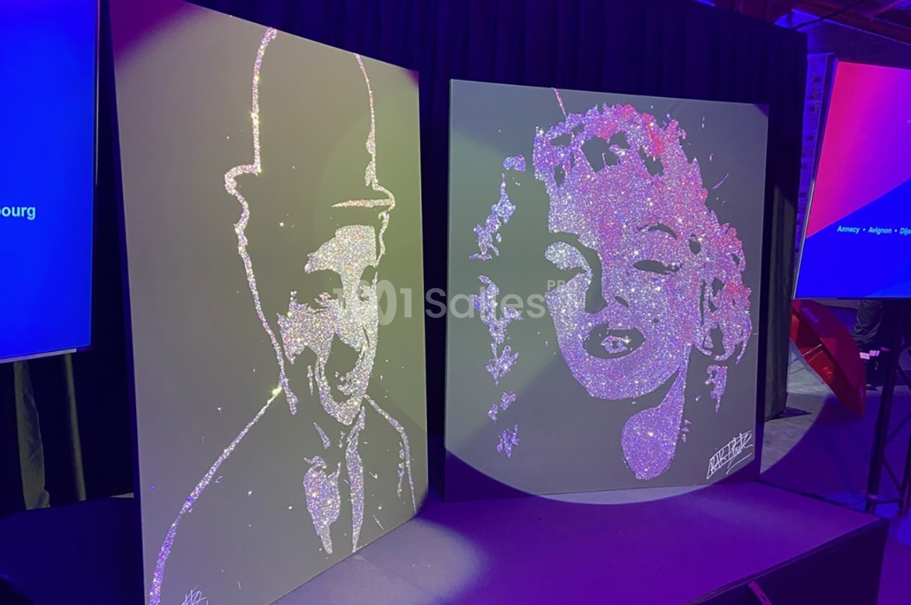 Portraits scintillants de deux figures célèbres réalisés en paillettes sur des panneaux noirs, exposés sur une scène.