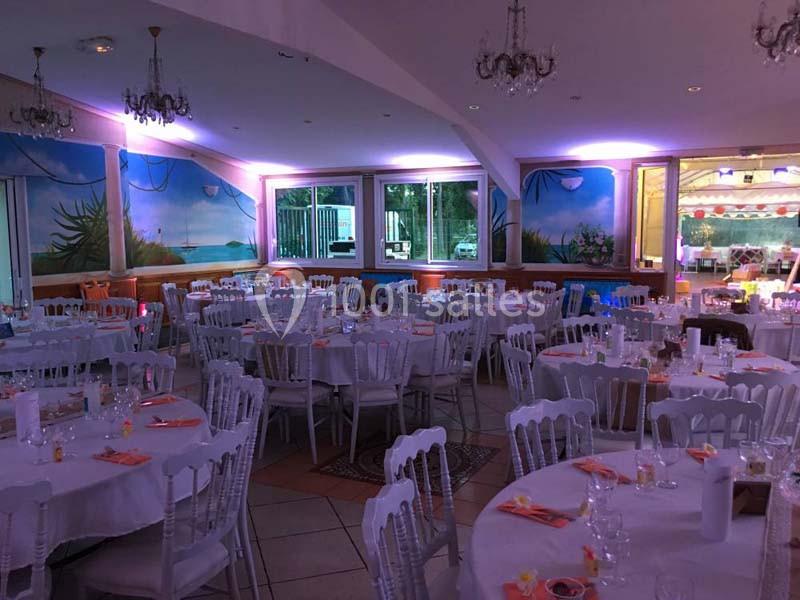 Salle de réception décorée avec des tables rondes, nappes blanches, chaises en bois et éclairage tamisé.