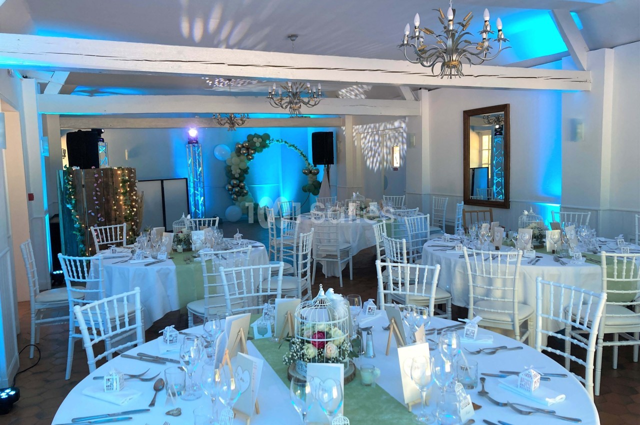 Salle de réception décorée pour un événement, avec tables rondes dressées, chaises blanches et éclairage bleu.