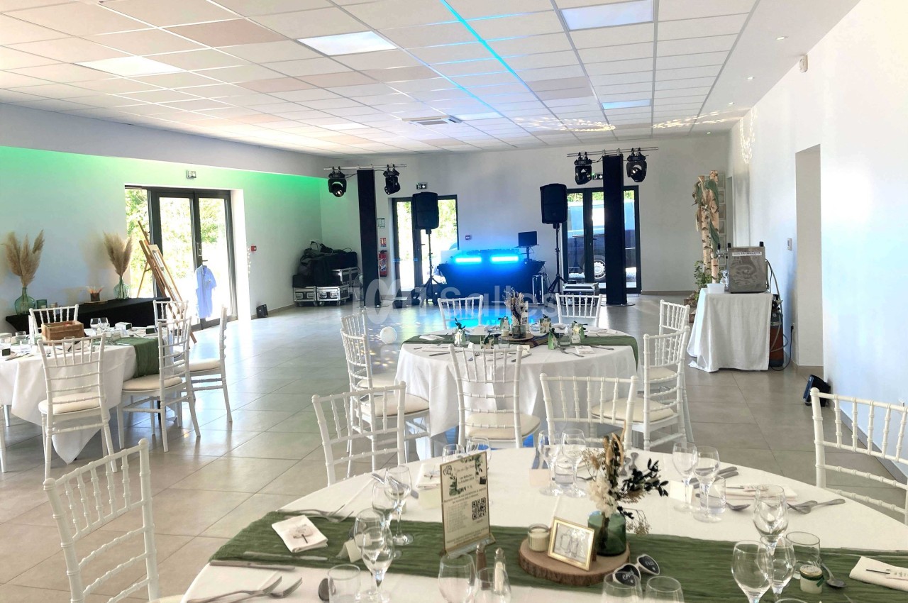 Salle de réception lumineuse avec tables dressées, chaises blanches et espace pour une piste de danse équipée de matériel…