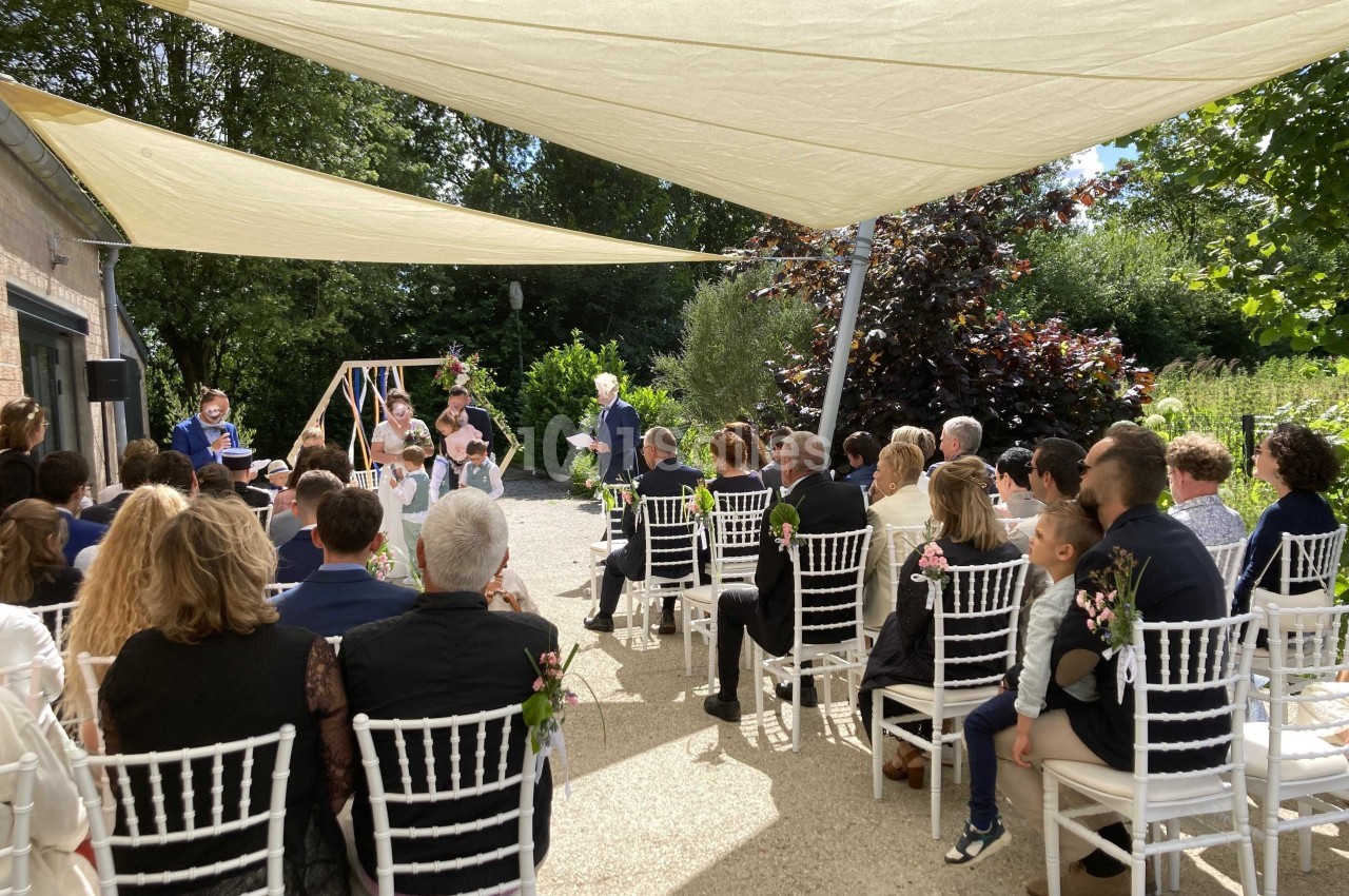 Cérémonie en plein air avec des invités assis sous des voiles d'ombrage, face à un officiant et un couple.
