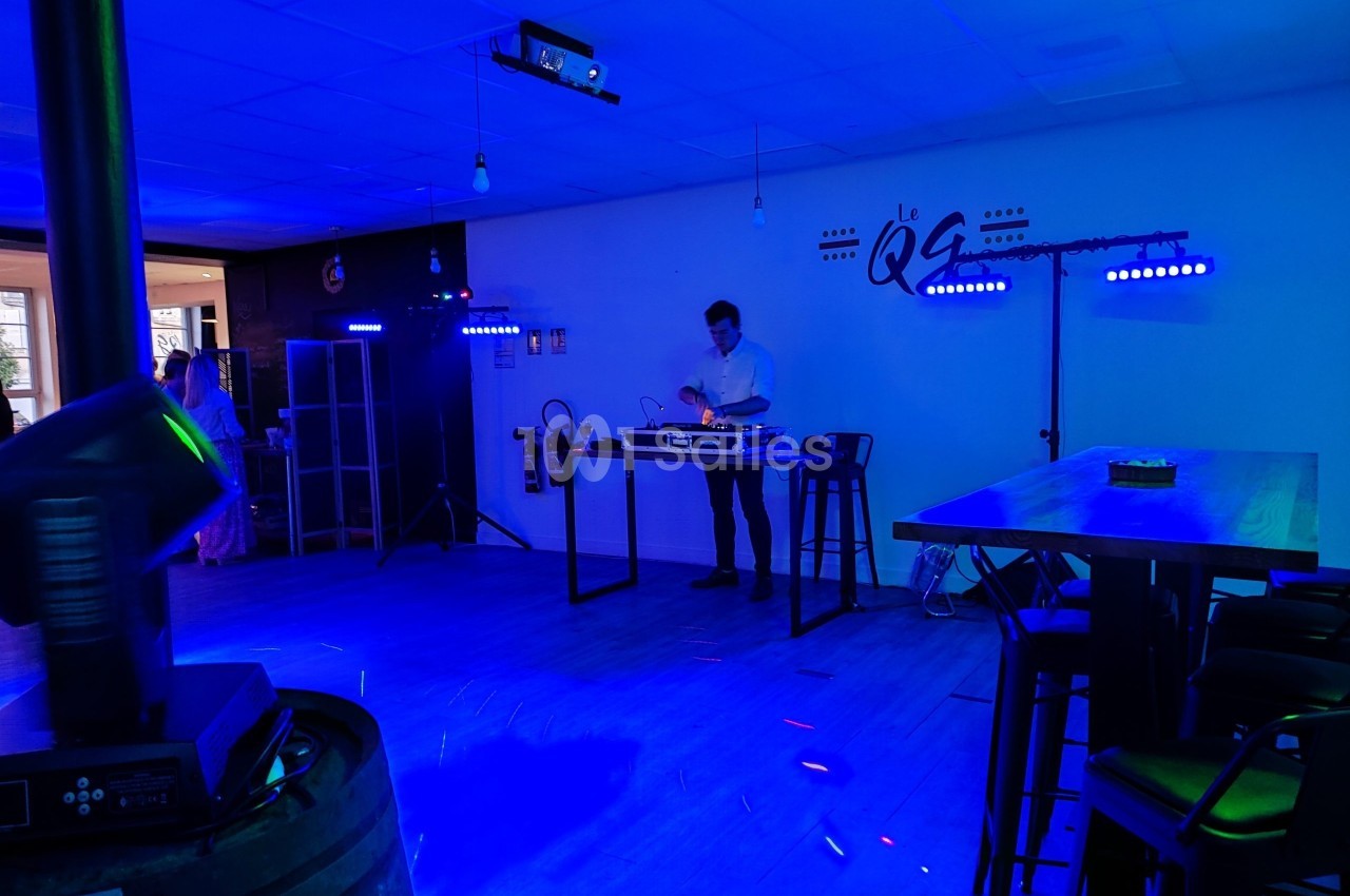Un DJ mixe dans une salle éclairée par des lumières colorées, avec des tables hautes et un décor sobre.