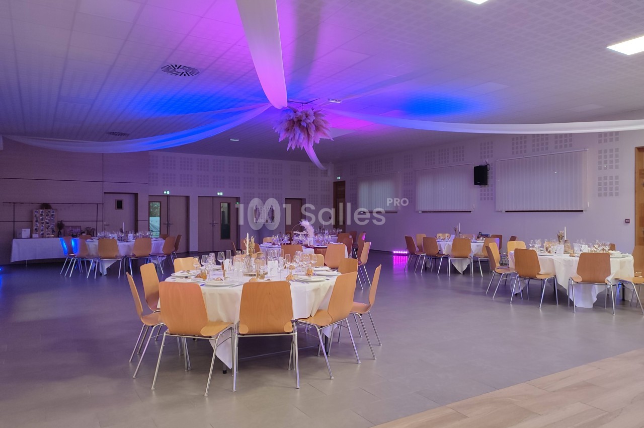 Salle de réception décorée avec des tables rondes dressées, éclairage violet et blanc, plafond orné de voilages.