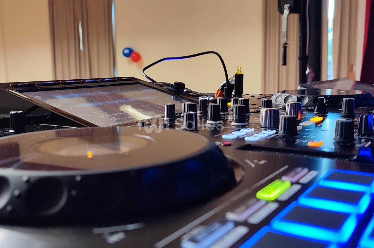 Table de mixage avec platine, boutons et écran tactile, installée dans une salle éclairée avec rideaux et ballons en arrière…