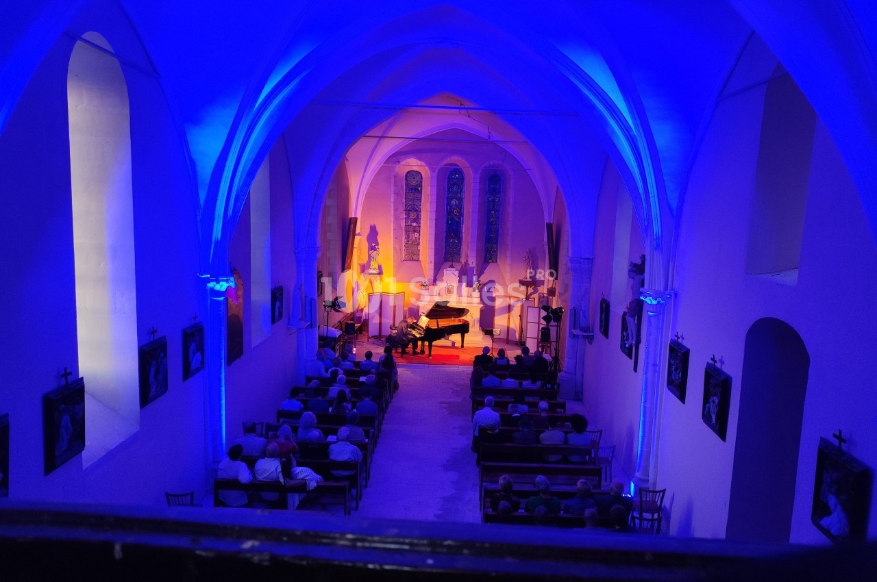 Intérieur d'une église illuminée en bleu et orange, avec un concert de piano devant un public assis.