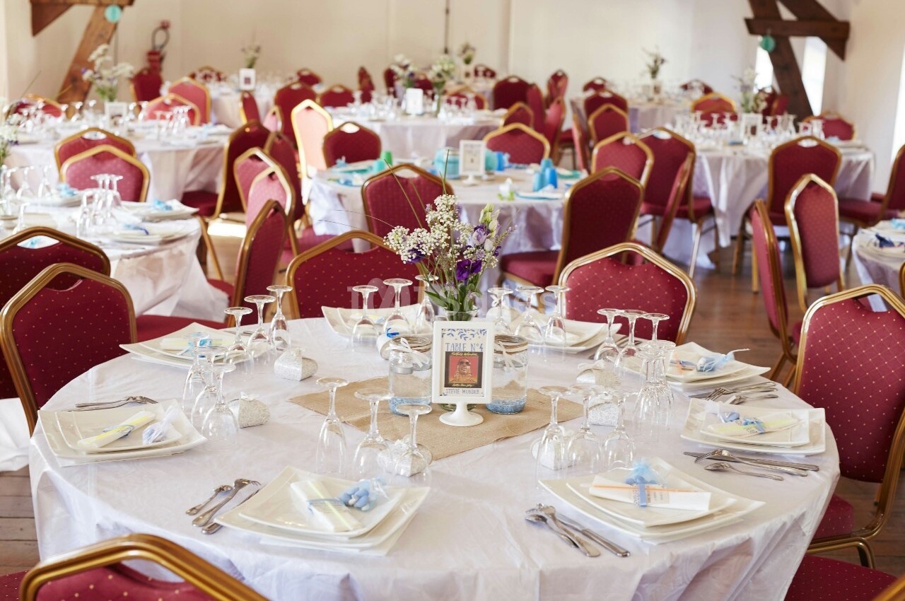 Salle de réception décorée avec des tables rondes dressées, nappes blanches, chaises rouges et centres de table fleuris.