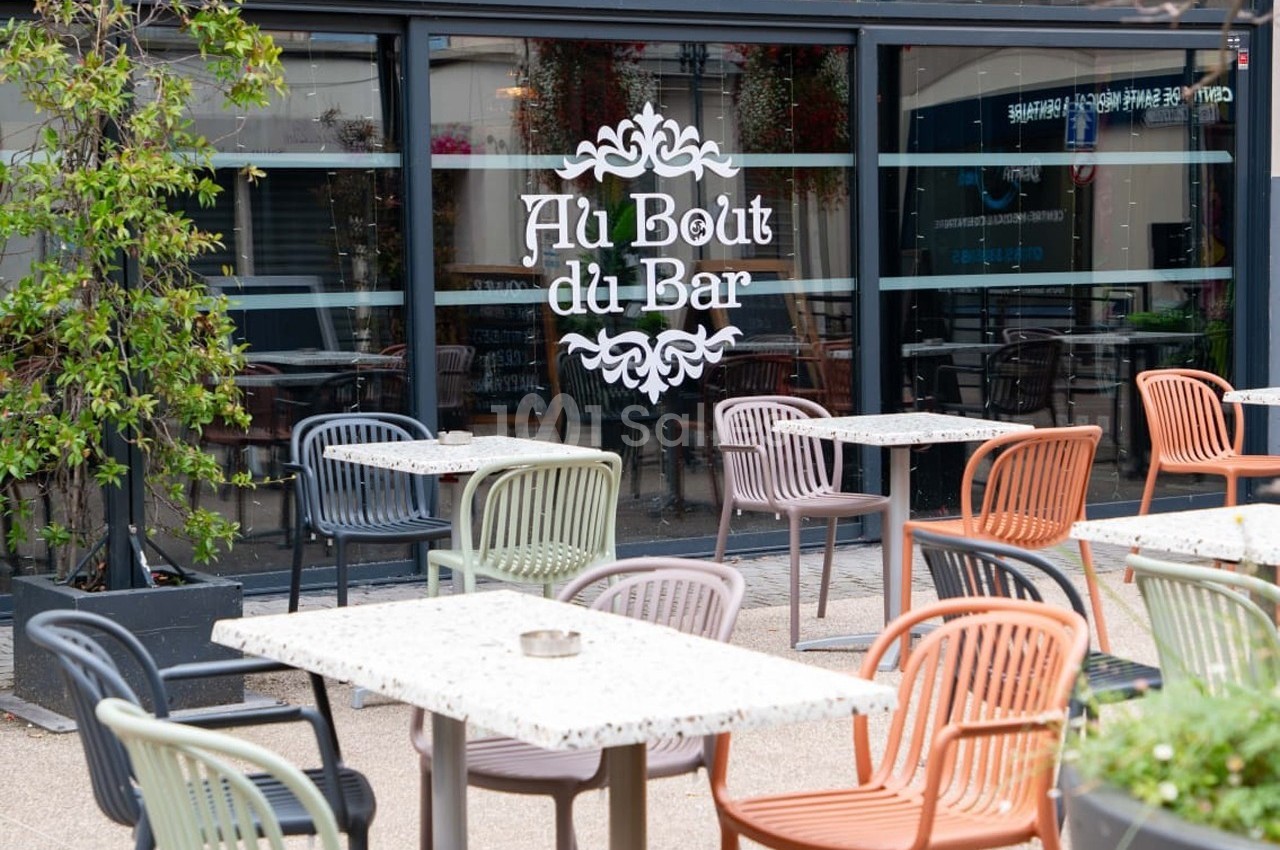 Terrasse d'un café avec des tables et des chaises colorées devant une façade vitrée portant l'inscription ’Au Bout du Bar’.