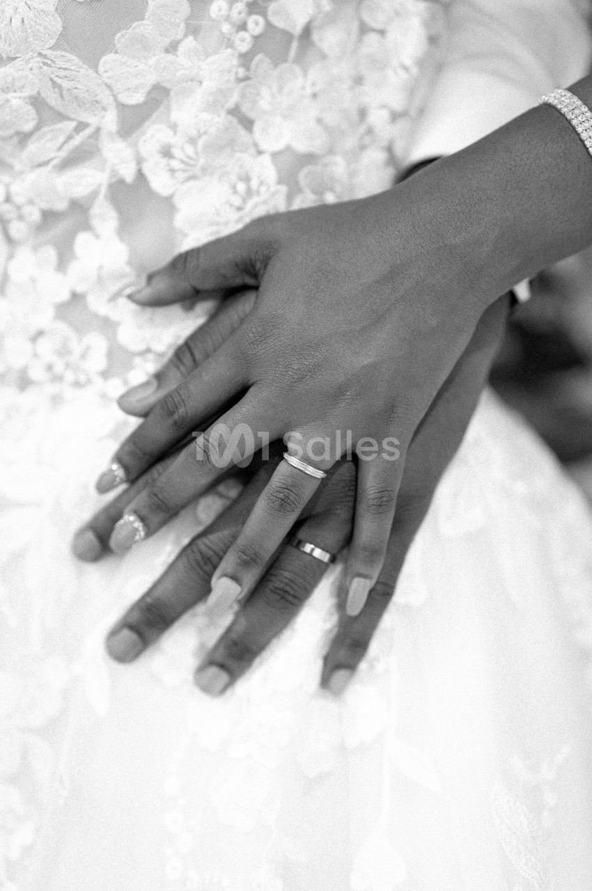 Mains d'un couple portant des alliances, posées l'une sur l'autre, sur une robe en dentelle blanche.