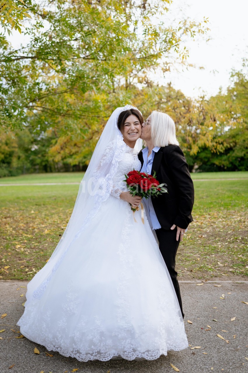 Une mariée souriante en robe blanche est embrassée sur la joue par une personne en costume noir dans un parc.