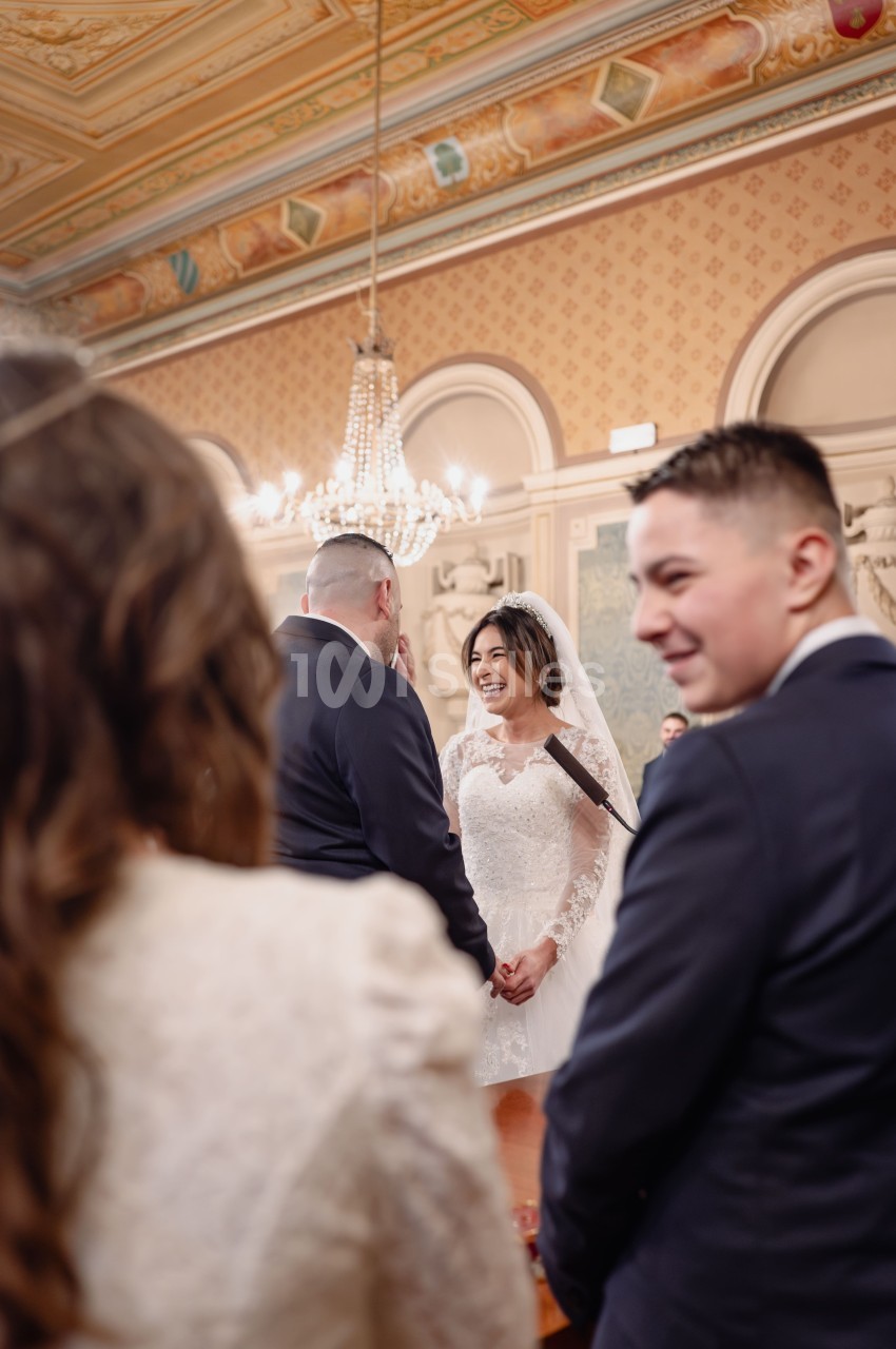 Un couple échange ses vœux de mariage dans une salle ornée de fresques et d'un lustre imposant.