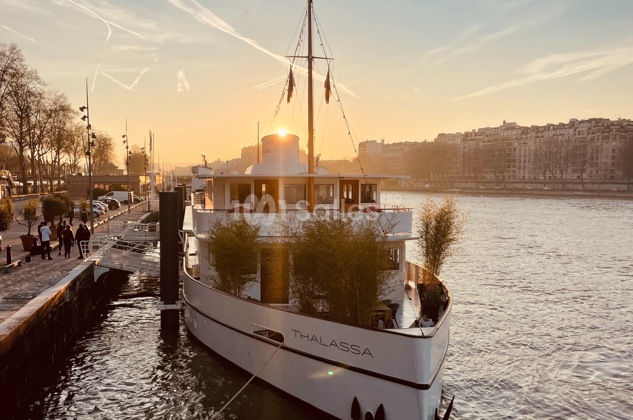 Bateau amarré sur une rivière au coucher du soleil, avec des arbres et des bâtiments en arrière-plan.