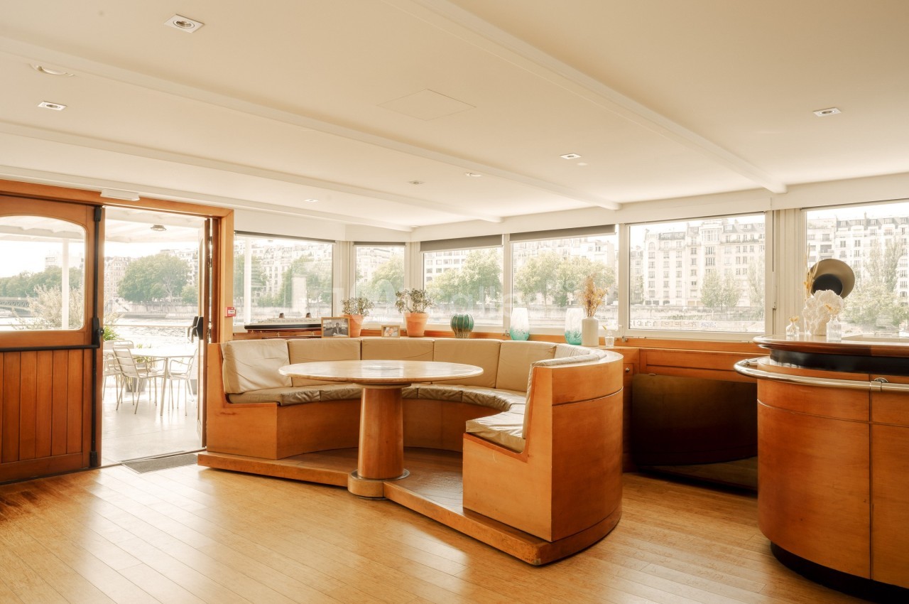 Intérieur lumineux d'une péniche avec banquettes en bois, table ronde et vue sur des bâtiments au bord de l'eau.
