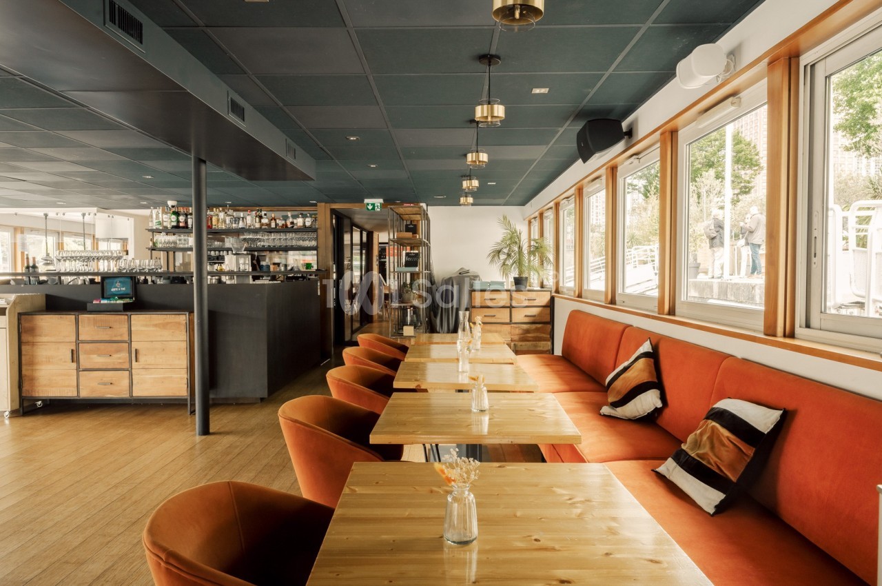 Salle de restaurant lumineuse avec tables en bois, chaises orange et grandes fenêtres donnant sur l'extérieur.