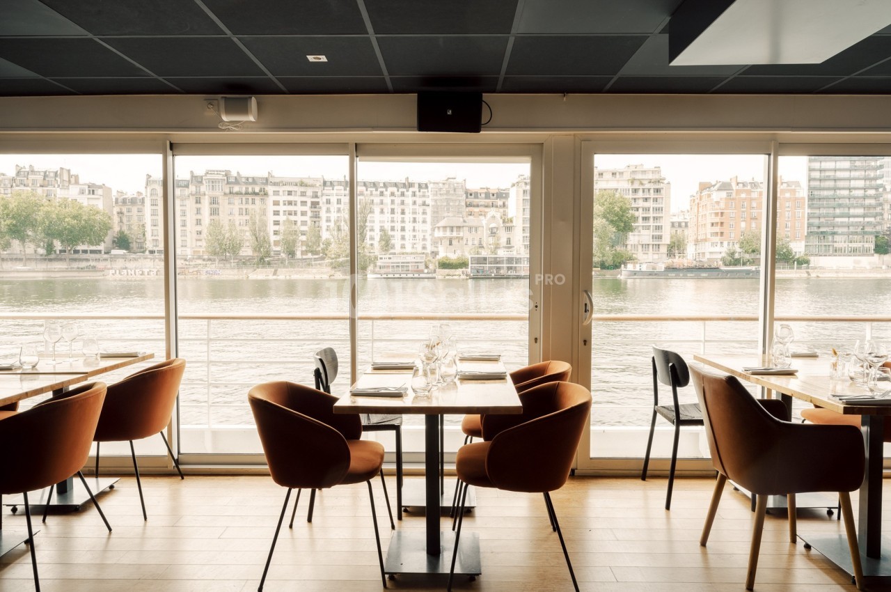 Salle de restaurant lumineuse avec des tables dressées, des chaises modernes et vue sur une rivière bordée de bâtiments.