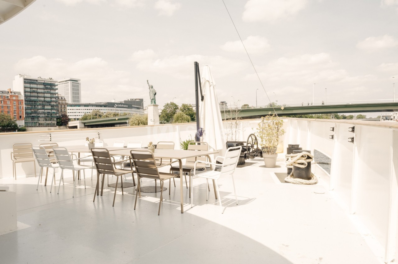 Terrasse extérieure d'un bateau avec des tables et chaises, vue sur un pont, des bâtiments et une statue au loin.