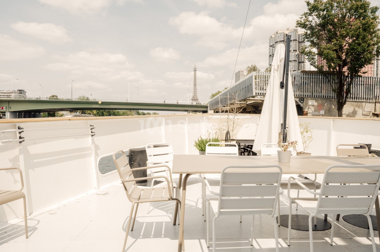 Terrasse extérieure d'un bateau avec table, chaises et parasol, vue sur un pont et la tour Eiffel en arrière-plan.