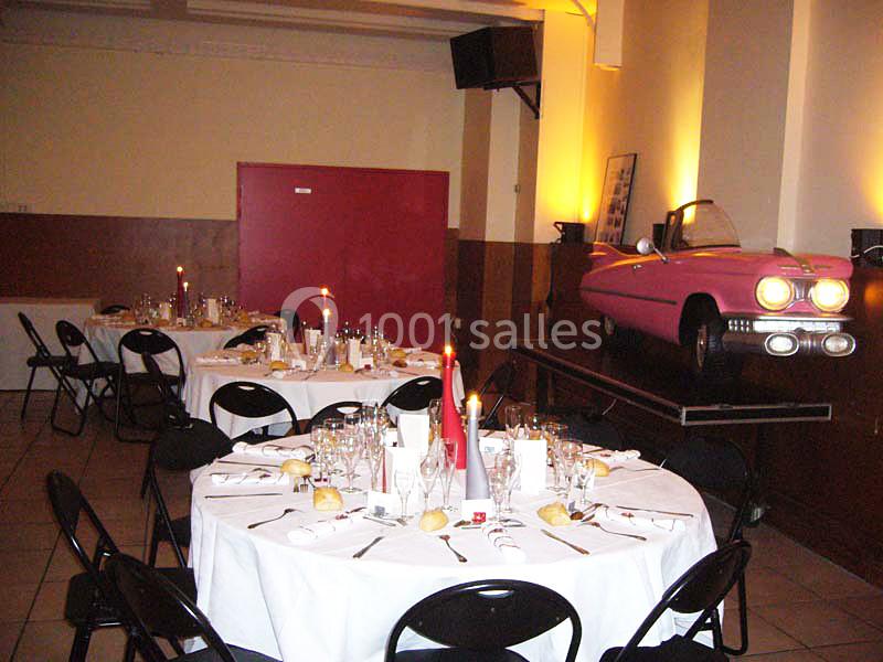 Salle de réception décorée avec des tables rondes dressées et une voiture rose exposée sur une estrade.