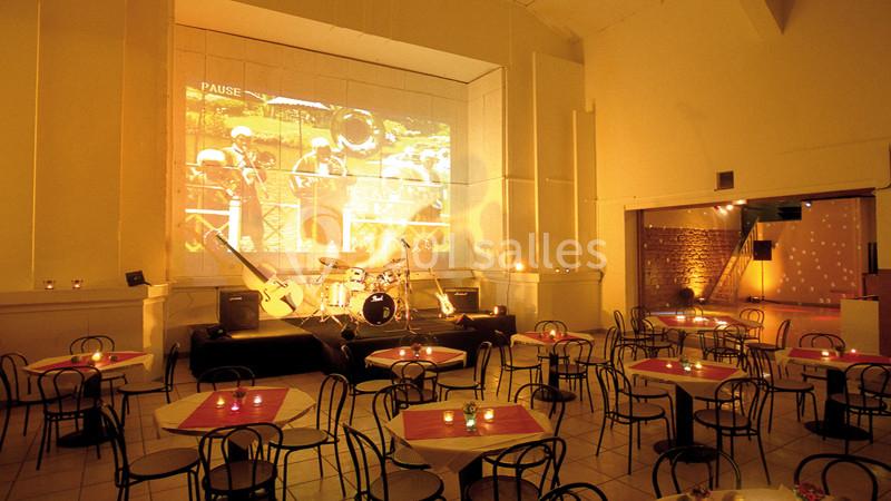 Salle de spectacle avec tables et chaises, petite scène équipée d'une batterie et projection murale en arrière-plan.