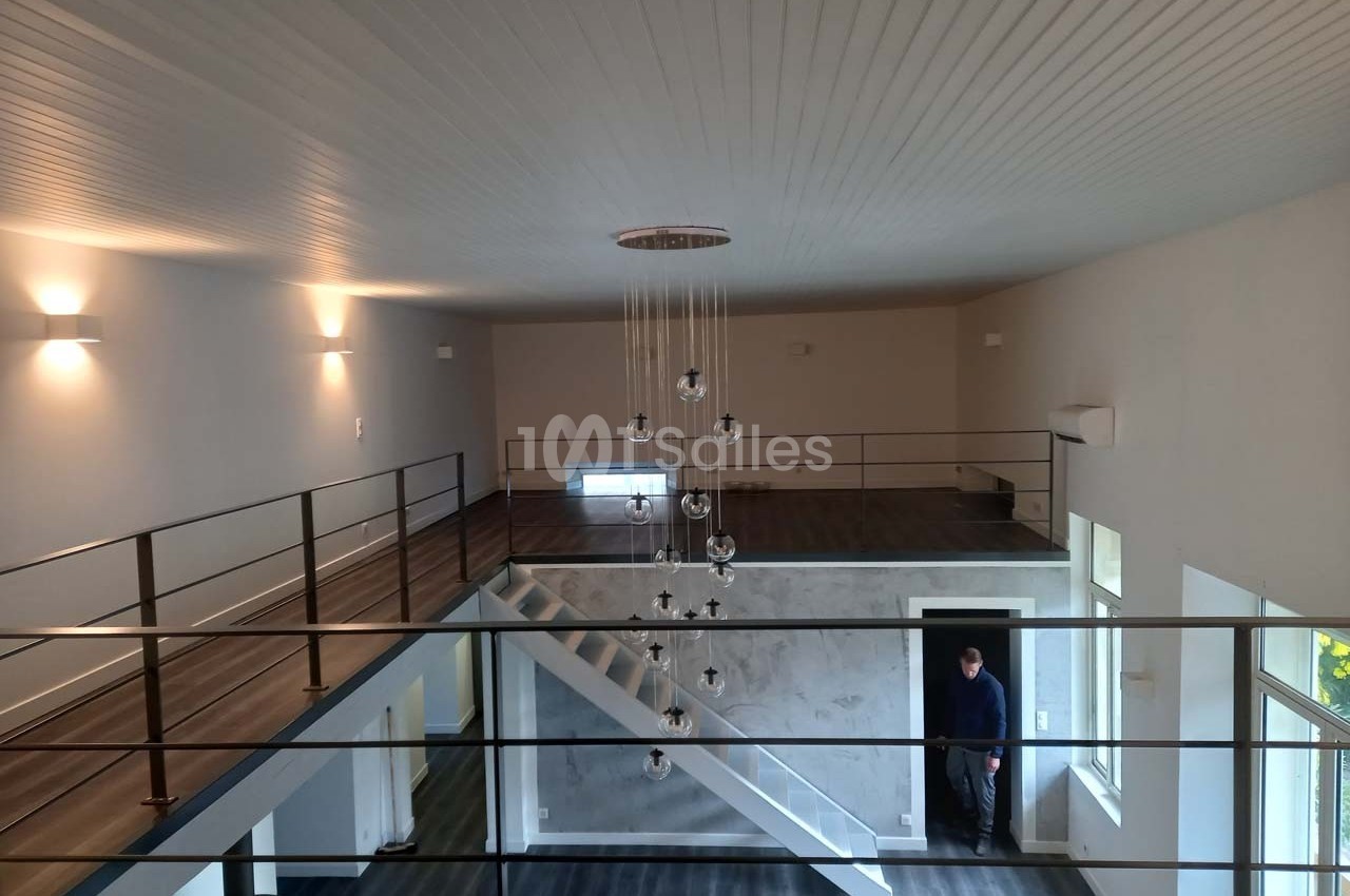 Vue intérieure d'un espace moderne à deux niveaux avec mezzanine, escalier central et éclairage suspendu.