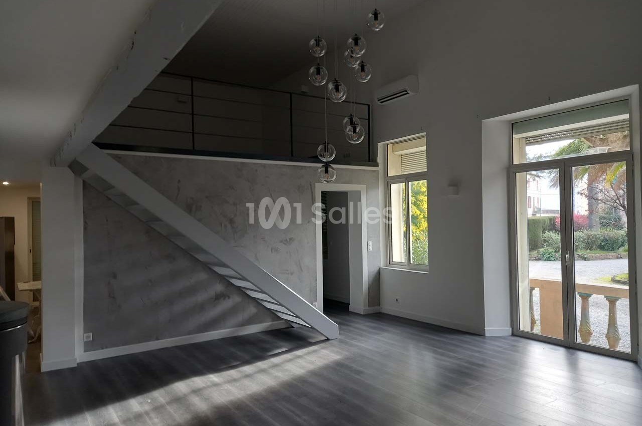 Intérieur lumineux avec mezzanine, escalier métallique, parquet sombre et grandes fenêtres donnant sur un jardin.