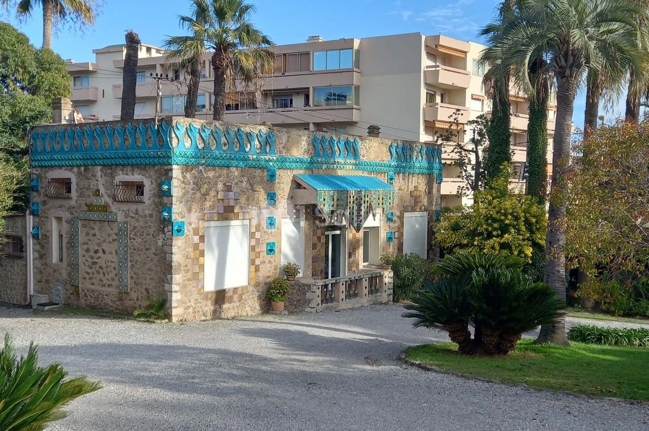 Maison en pierre ornée de mosaïques bleues, entourée de palmiers et de végétation, avec des immeubles en arrière-plan.