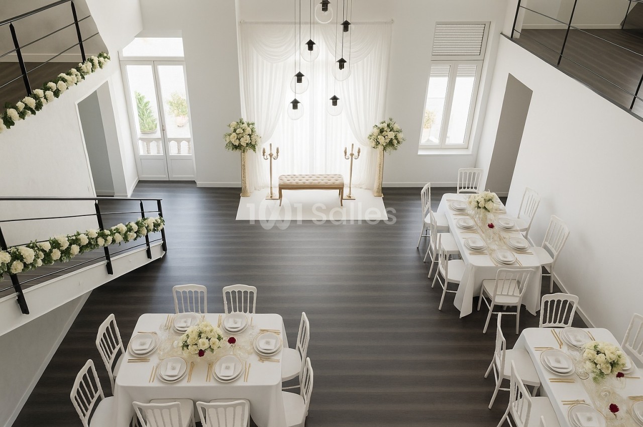Salle lumineuse décorée pour un mariage, avec tables dressées, fleurs blanches et espace pour une cérémonie.
