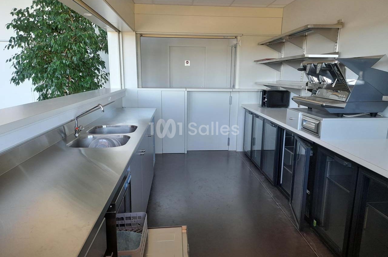 Cuisine professionnelle avec plan de travail en inox, évier double, étagères murales, machines à café et réfrigérateurs.