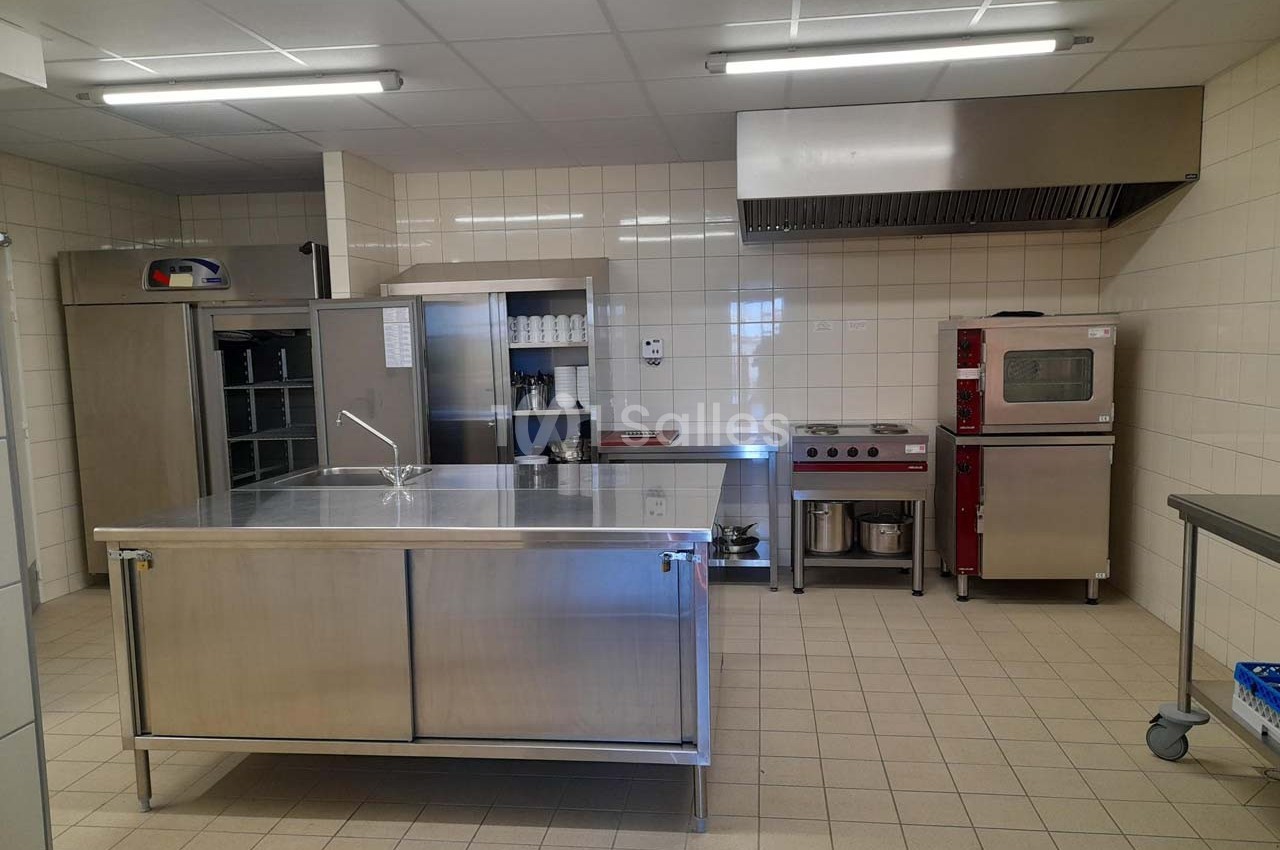 Cuisine professionnelle avec plan de travail en inox, équipements de cuisson, four, étagères et carrelage mural beige.