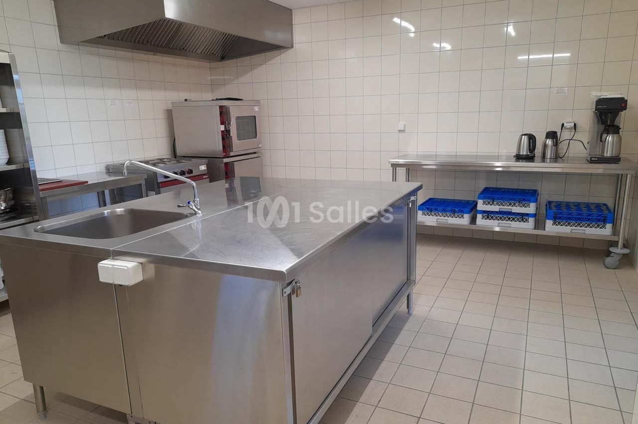 Cuisine professionnelle avec plan de travail en inox, évier, étagères, four, cafetière et carrelage mural blanc.