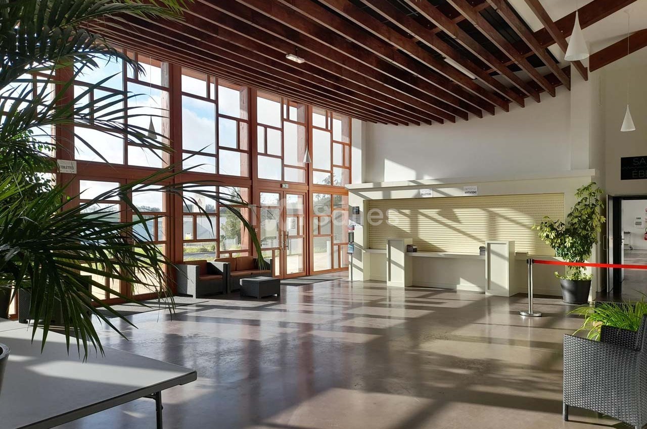 Hall lumineux avec de grandes baies vitrées, des plantes, un comptoir d'accueil et un sol en béton poli.