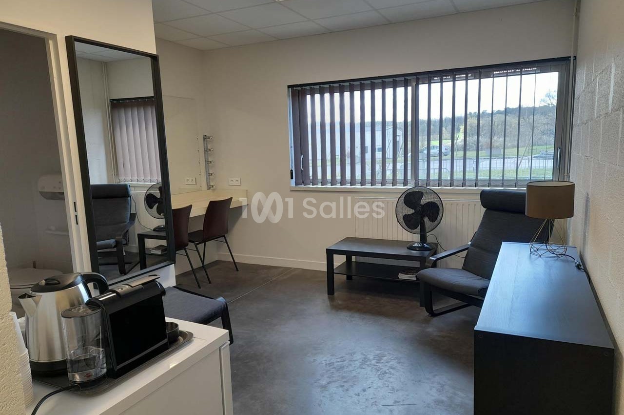 Salle lumineuse avec bureau, fauteuil, miroir, cafetière et fenêtre à stores donnant sur un extérieur verdoyant.