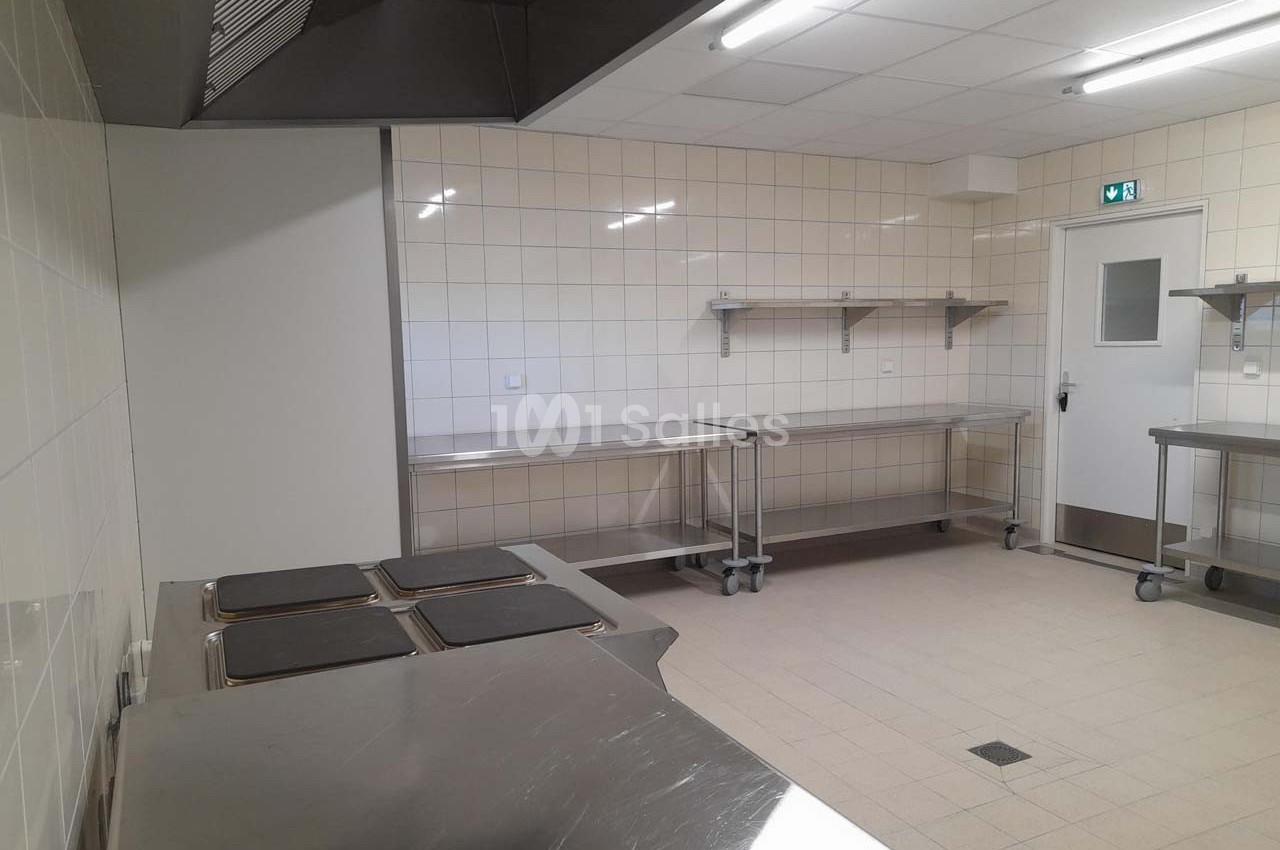 Cuisine professionnelle vide avec plans de travail en inox, carrelage mural blanc et éclairage fluorescent.