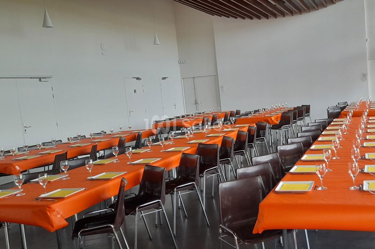 Salle aménagée avec des tables rectangulaires couvertes de nappes orange, disposées en rangées, avec chaises et verres.