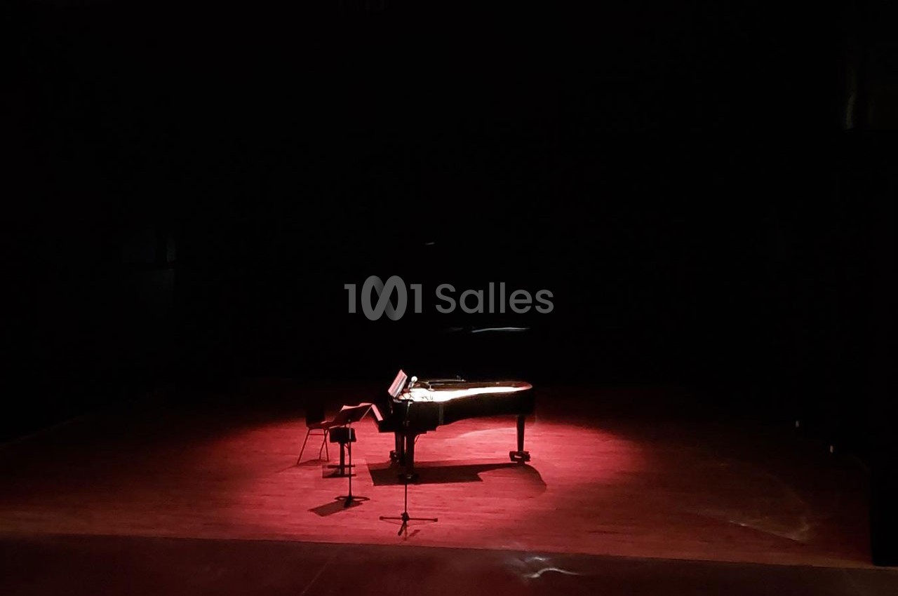 Un piano à queue éclairé par une lumière rouge sur une scène sombre, avec un tabouret et un pupitre.