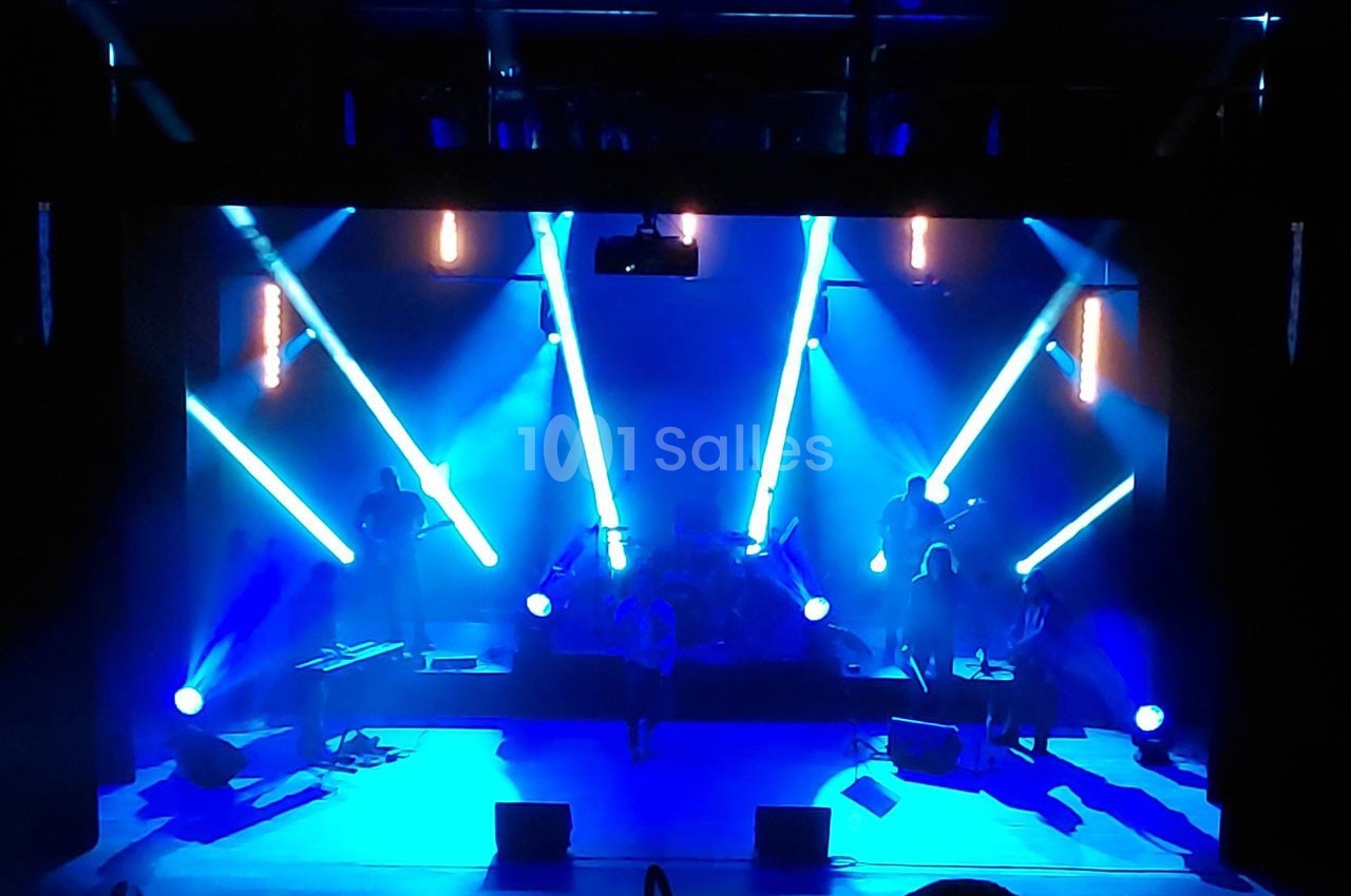 Concert sur scène avec éclairage bleu intense, musiciens en silhouette et projecteurs lumineux dirigés vers le public.