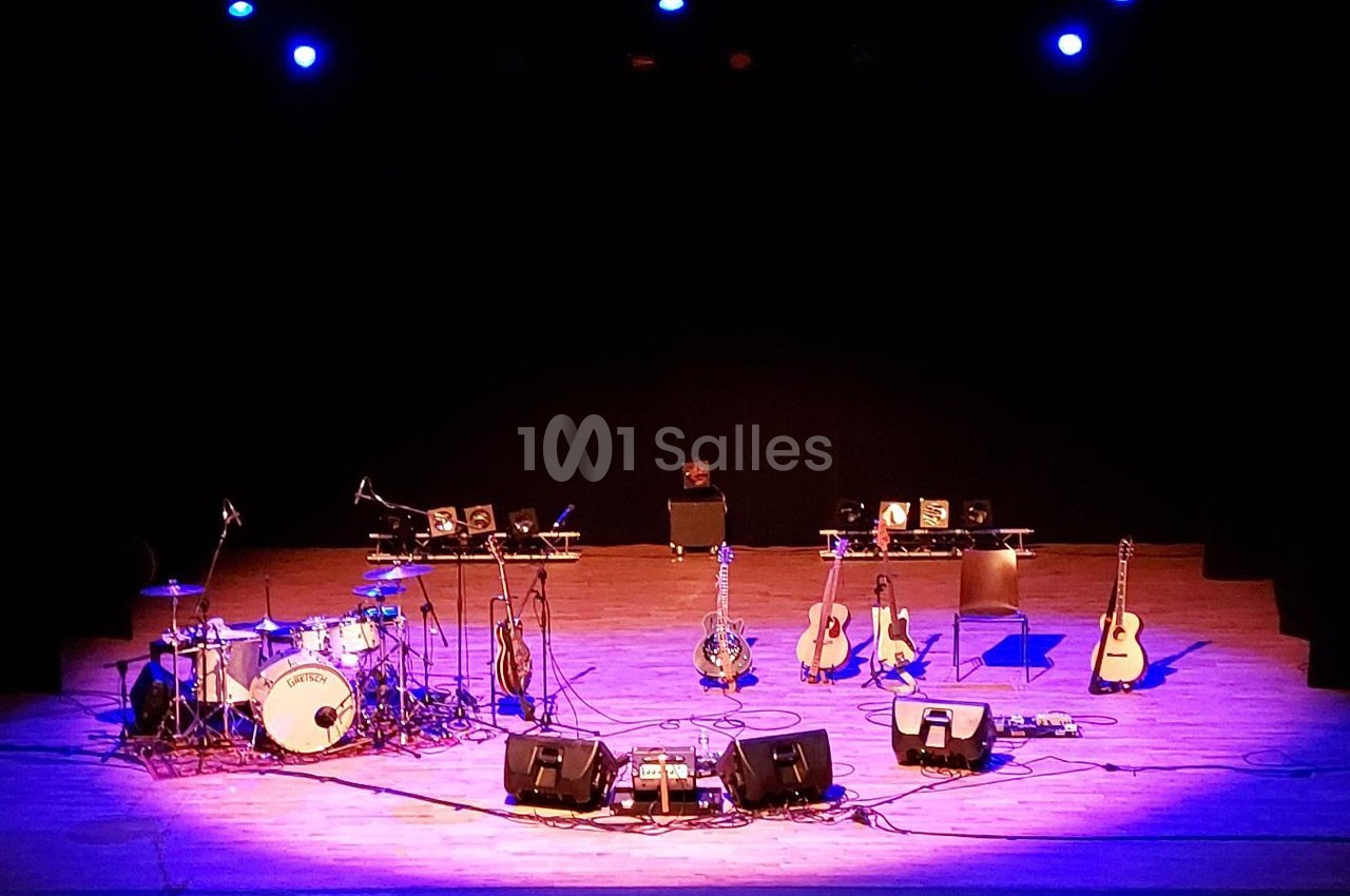 Scène de concert vide avec des instruments de musique, dont des guitares, une batterie et des micros, sous un éclairage bleu.