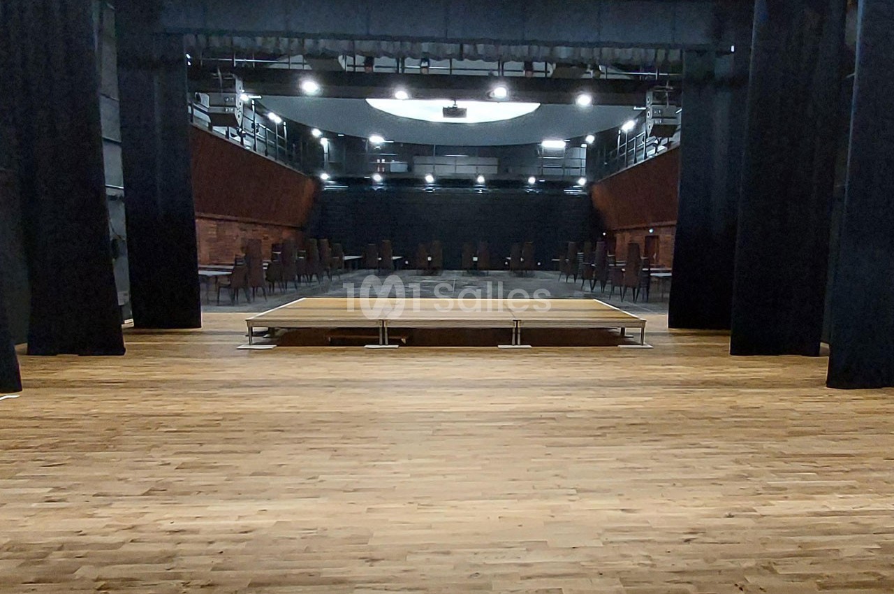Salle de spectacle vide avec une scène en bois au centre, entourée de rideaux noirs et de rangées de sièges.