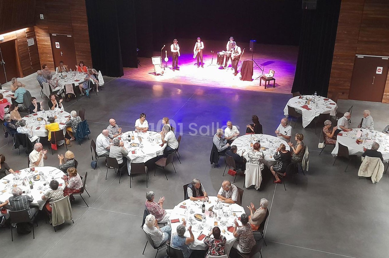 Des personnes assises autour de tables rondes dans une salle, assistent à une performance musicale sur scène.