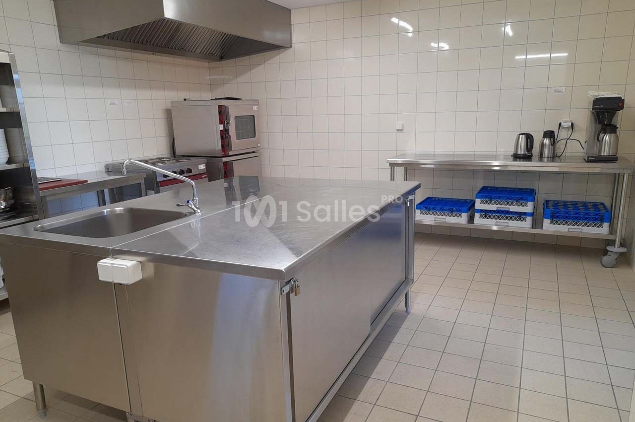 Cuisine professionnelle avec plan de travail en inox, évier, équipements de cuisson et étagères de rangement.