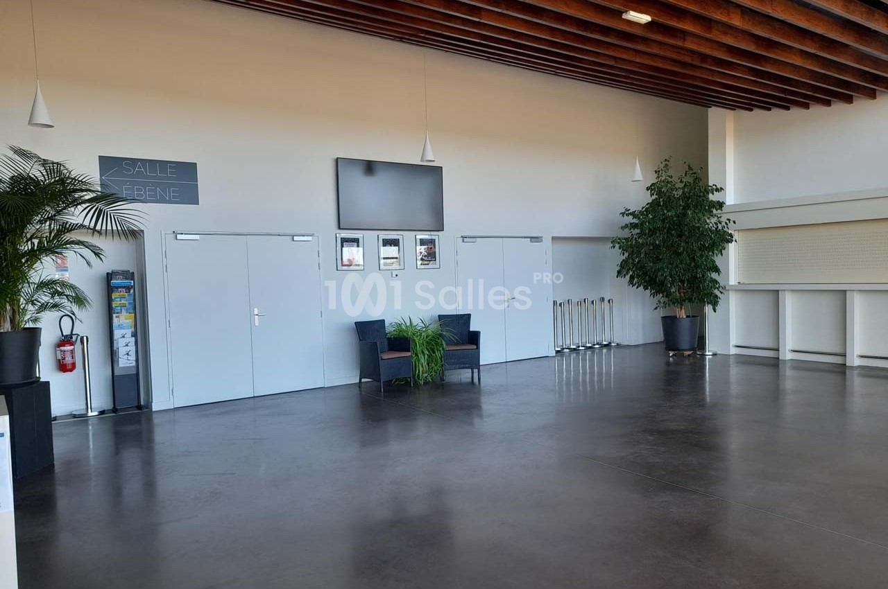 Hall lumineux avec sol en béton, plantes en pot, chaises, panneaux muraux et plafond en bois avec suspensions.