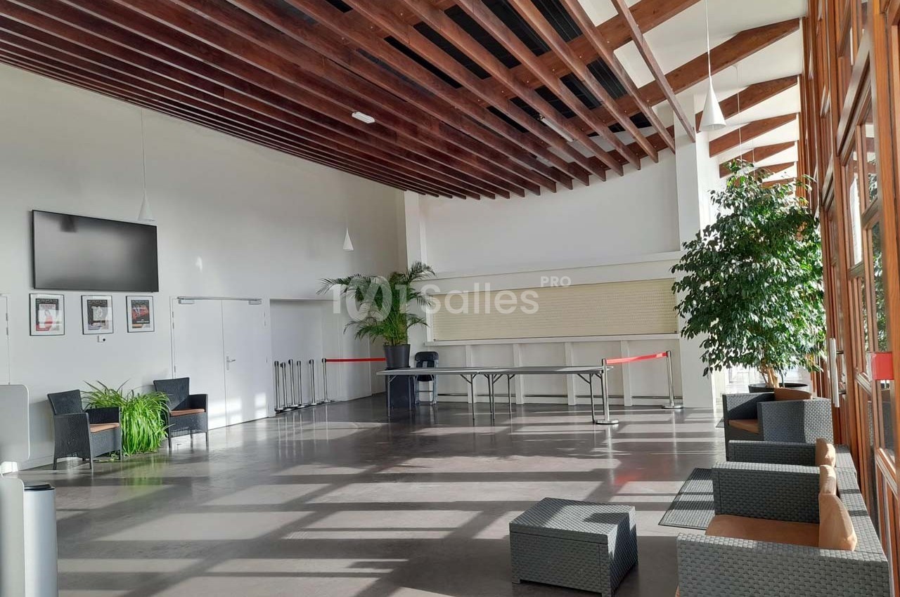 Hall lumineux avec grandes baies vitrées, mobilier moderne, plantes vertes et plafond en bois.