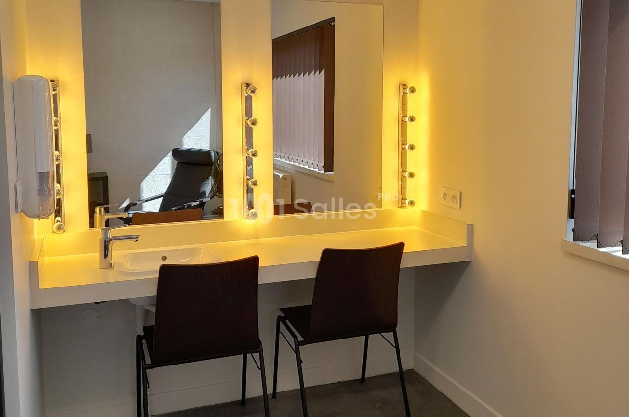 Coiffeuse avec miroir éclairé par des ampoules, deux chaises noires et un lavabo intégré dans une pièce lumineuse.
